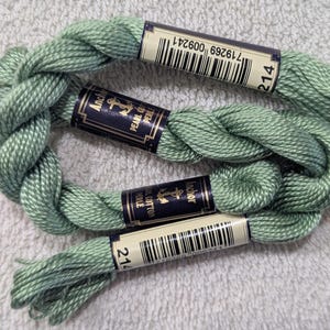 May include: Three skeins of Anchor Pearl Cotton thread in a soft, muted green colour. Each skein is labelled with a barcode and the number 214. The thread is tightly wound and displayed on a textured, white surface.
