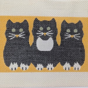 May include: Needlepoint canvas featuring three black cats with white whiskers and green eyes, set against a yellow background. The cats have white paws and are on a cream-colored canvas.