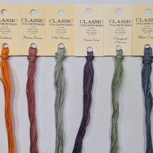 May include: A collection of Classic Colorworks embroidery floss in various colors, including green, purple, and brown. Each skein is labeled with its color name, such as "Frog Legs" and "Blue Corn."