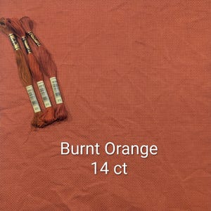May include: Burnt orange 14 ct cross stitch fabric with three skeins of embroidery floss in shades of brown and orange. The fabric is a solid color with a woven texture. The text "Burnt Orange 14 ct" is displayed in white.