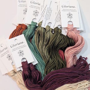 May include: A collection of Gloriana hand-dyed ribbons and threads for fine needlework, featuring various colors including cream, rust, teal, olive green, pink, and deep purple, each attached to a white card with the brand name and logo.