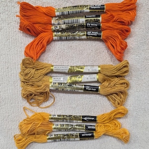 Könnte beinhalten: Eine Auswahl an sechssträngigem Stickgarn in Orange- und Goldtönen. Die Farben sind mit Namen wie "Tangerine" und "Bright Gold" gekennzeichnet. Jedes Knäuel ist etwa 8,2 Meter lang.