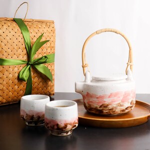 Peut inclure: Une théière en céramique avec une anse en rotin courbée et deux tasses assorties, toutes dotées d'une base blanche avec des motifs de coups de pinceau roses et bruns. L'ensemble repose sur un plateau en bois rond près d'un panier tressé carré.