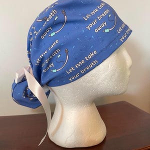 May include: Blue surgical scrub hat with a white ribbon tie. The hat has a repeating pattern of a medical breathing tube and the text "Let me take your breath away".