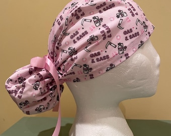 Può includere: Un berretto chirurgico rosa con una cravatta. Il berretto ha un motivo ripetuto di un serbatoio di gas, una siringa, un bisturi e il testo "Gas & Sass".