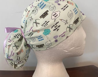 May include: A light green scrub cap with a ponytail, adorned with medical-themed illustrations and phrases like "In your wildest dreams" and "Meet Me at the Afterglow." The cap is tied with a lavender ribbon.