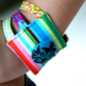 May include: A colorful fabric bracelet with a black button featuring a floral design. The bracelet is made of multiple strips of fabric sewn together, creating a layered effect. The fabric is striped with pink, blue, green, and yellow.