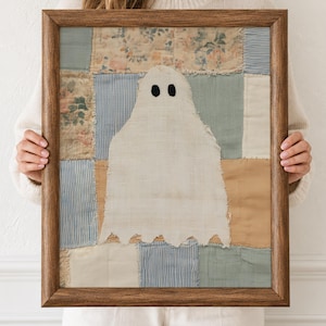 Könnte beinhalten: Ein gerahmtes Textilkunstwerk mit einem zentralen weißen Geister-Applikat mit zwei schwarzen runden Augen vor einem Patchwork-Hintergrund aus verschiedenen Stoffquadraten in Blau-, Beige- und Blumenmustern in einem braunen Holzrahmen.