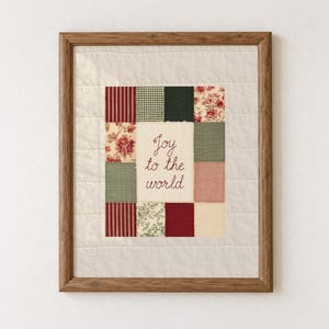 May include: A framed wall art piece featuring a central cream-colored fabric panel with the embroidered text Joy to the world, surrounded by a patchwork border of various red, green, and floral patterned fabric squares, all set in a wood frame.
