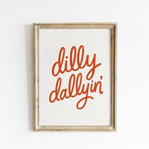 May include: A framed print with the words "dilly dallyin'" in red script on a white background. The frame is a light, neutral color, and the print is centered. The overall aesthetic is simple and modern.
