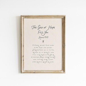 Peut inclure: Une impression encadrée avec le texte "THE GOD OF HOPE FILL YOU" et la référence biblique Romains 15:13. L'impression a un fond crème et est dans un cadre en bois brun clair. Le texte est en noir.