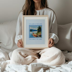 May include: A framed print with the text "THIS LAND IS YOUR LAND" and "THIS LAND IS MY LAND". The artwork depicts a boat on a lake with mountains in the background. The frame is a light wood color.