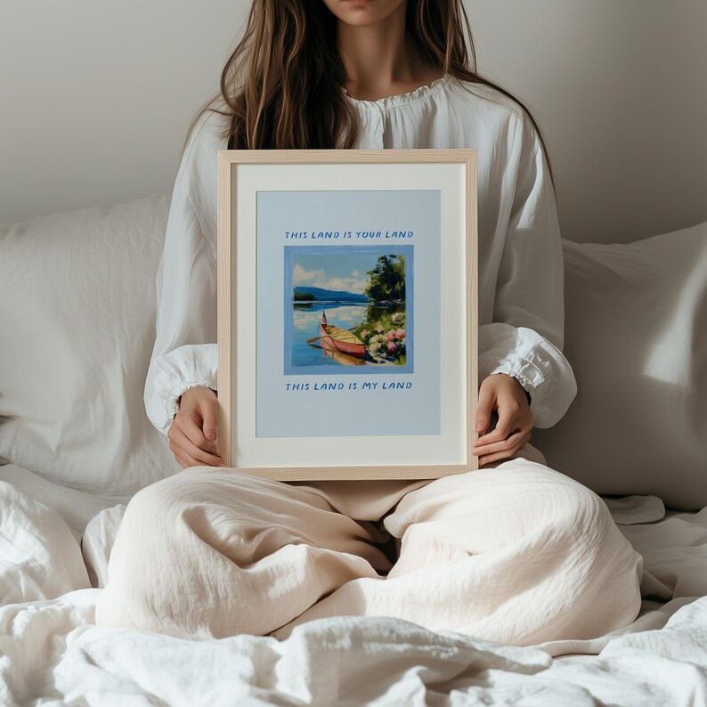 May include: A framed print with the text "THIS LAND IS YOUR LAND" and "THIS LAND IS MY LAND". The artwork depicts a boat on a lake with mountains in the background. The frame is a light wood color.