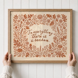 May include: A framed print featuring the text "To everything there is a season" in brown script, surrounded by a detailed botanical border of rust-colored oak leaves, acorns, wheat stalks, pumpkins, and flowers on a light cream background.