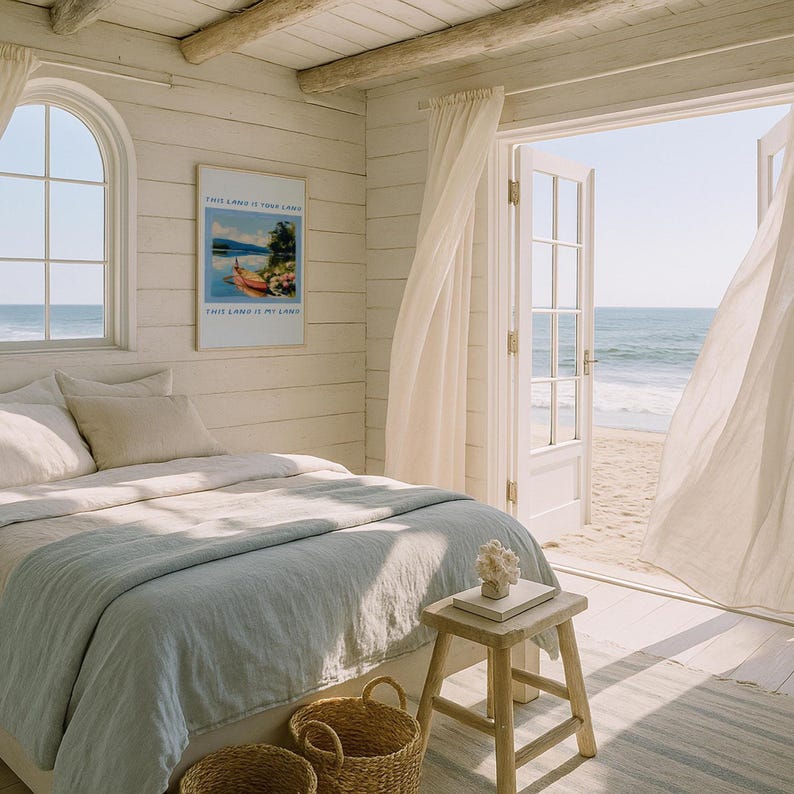May include: A bright bedroom with a view of the ocean. The room features a bed with a light blue blanket, a small wooden stool, and a framed poster on the wall. The open doors reveal a sandy beach and the sea. The text on the poster reads "THIS LAND IS YOUR LAND".