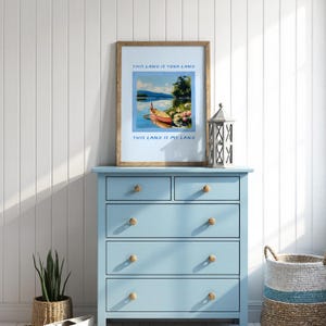 May include: A framed art print with the text "THIS LAND IS YOUR LAND" and "THIS LAND IS MY LAND" above and below a landscape painting of a boat on a lake. The print is displayed on a light blue dresser with wooden knobs.