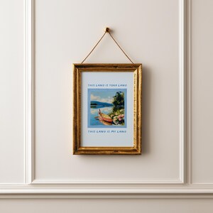May include: A framed print with a gold-colored frame hanging on a white wall. The print features a landscape with a boat on a lake, with the text "THIS LAND IS YOUR LAND" and "THIS LAND IS MY LAND".