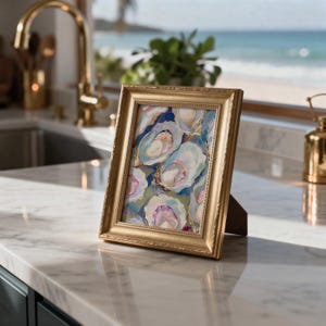 May include: A framed artwork featuring an oyster painting. The gold-colored frame surrounds a painting of several oysters in shades of blue, pink, and white. The artwork is displayed on a marble countertop, with a beach scene visible in the background.