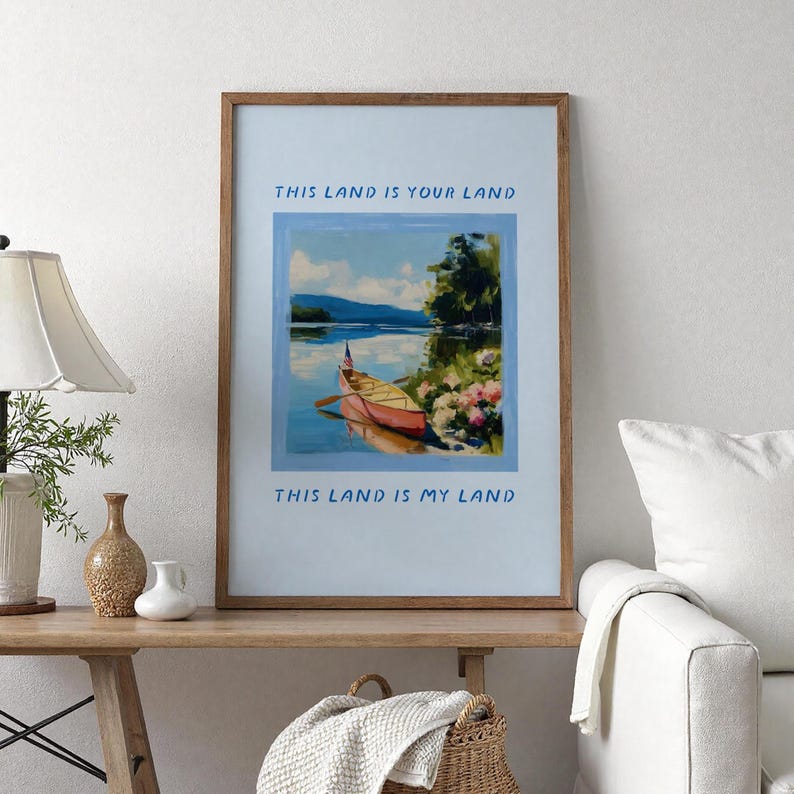 May include: A framed art print featuring a landscape painting of a lake with a canoe and the text "THIS LAND IS YOUR LAND" and "THIS LAND IS MY LAND". The artwork is in a wooden frame and is displayed on a wooden table.