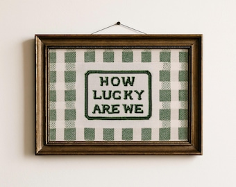 May include: A framed cross-stitch artwork with the text "HOW LUCKY ARE WE" in green thread. The artwork is set against a green and white gingham pattern and framed in a dark wood frame. The piece is hanging on a white wall.
