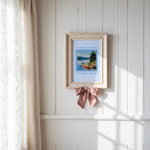 May include: A framed landscape print with the text "THIS LAND IS YOUR LAND" and "THIS LAND IS MY LAND". The frame is a distressed, light-colored wood. A dusty rose bow is tied below the frame. The print depicts a boat on a lake.