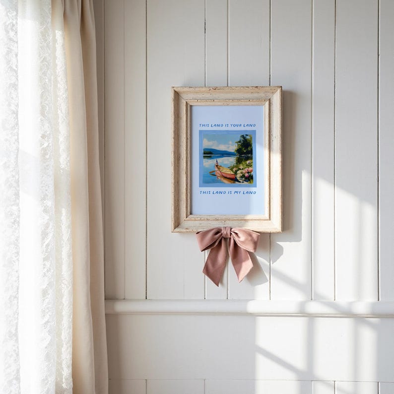 May include: A framed landscape print with the text "THIS LAND IS YOUR LAND" and "THIS LAND IS MY LAND". The frame is a distressed, light-colored wood. A dusty rose bow is tied below the frame. The print depicts a boat on a lake.
