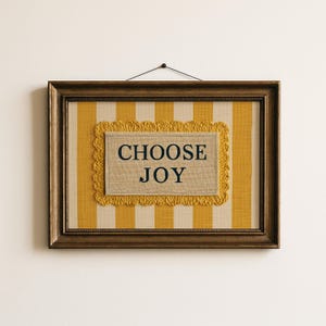 以下が含まれることがあります： 金色のフレームに入った壁掛けアート。 黄色とクリーム色のストライプの背景に、中央に「CHOOSE JOY」と紺色の文字で書かれた長方形のパッチがあります。 パッチは黄色の装飾的なボーダーで囲まれています。
