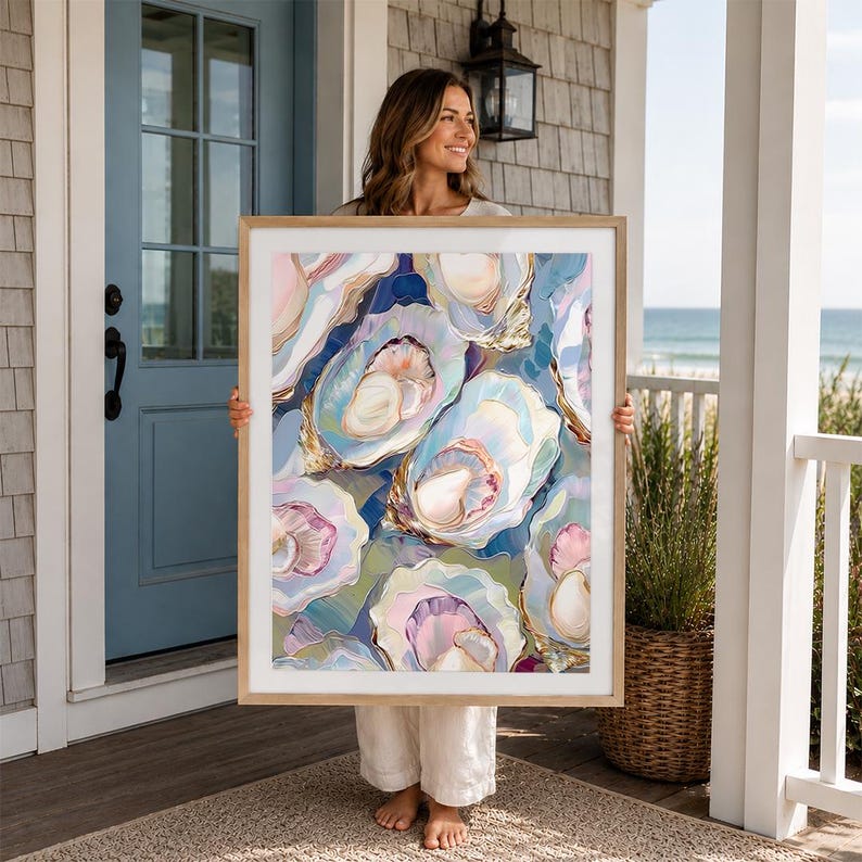 May include: A framed art print featuring an abstract painting of oysters in shades of blue, pink, and gold. The artwork has a light wood frame and is held by a person standing on a porch.