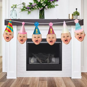 May include: A decorative party banner featuring six cutouts of smiling baby faces, each wearing a different coloured party hat. The hats include a rainbow striped cone, a pink striped cone, a blue glittery cone with the number 1, a pink glittery cone with the number 1, a plain light-coloured cone, and a blue and pink striped cone with a pom-pom.