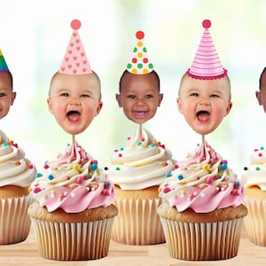 May include: Five cupcakes with white icing and colourful sprinkles topped with a baby face and a colourful party hat. The hats are rainbow, pink with hearts, red and green polka dots, pink and white stripes, and blue and white polka dots.
