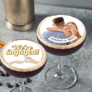 May include: Two cocktail glasses filled with a dark liquid, possibly coffee-based. The drinks are topped with circular designs. One reads "We're Engaged!" with a hand graphic. The other displays a photo of a couple with the text "Gemma & Ben".