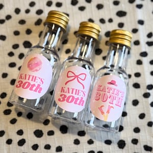 May include: Three clear glass bottles with pink and white labels that say "Katie's 30th". Each bottle has a different design on the label: a disco ball, a bow, and a cowboy hat.
