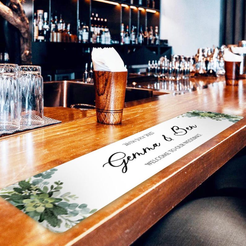 K&ouml;nnte beinhalten: Ein wei&szlig;er Bartischl&auml;ufer mit dem Text "Gemma & Ben" und "Willkommen zu unserer Hochzeit" in schwarzer Schrift. Der L&auml;ufer hat ein Blumendesign mit gr&uuml;nen Bl&auml;ttern und liegt auf einer Holzbar.