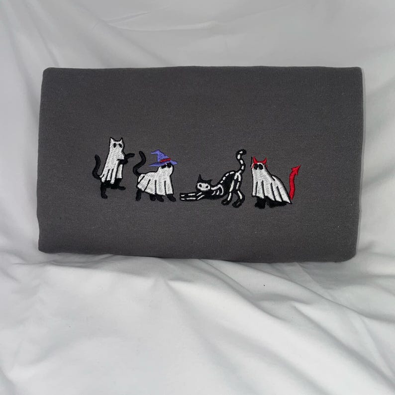 May include: A gray sweatshirt with an embroidered design of five cats dressed as ghosts for Halloween. One cat is wearing a witch's hat, another has a devil's tail, and the other three are wearing simple ghost costumes.