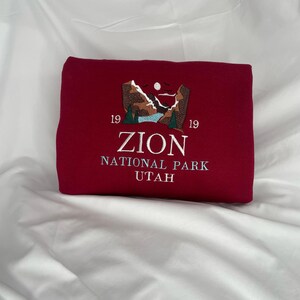 Pode incluir: Uma camisola cor de vinho com um desenho bordado de uma cordilheira com um lago e o texto "Zion National Park Utah" e "19 19" de cada lado do texto.