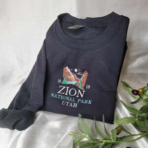Pode incluir: Camisola de moletom preta com um desenho bordado de uma cordilheira, um lago e uma lua. O texto "Zion National Park Utah" est&aacute; bordado abaixo do desenho.
