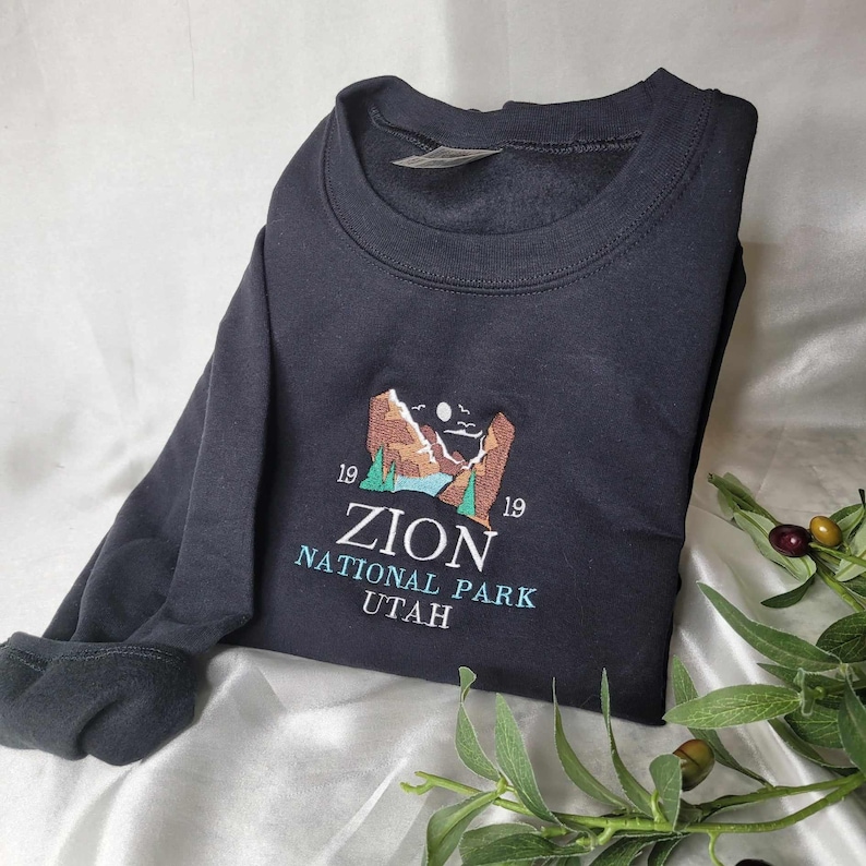 Pode incluir: Camisola de moletom preta com um desenho bordado de uma cordilheira, um lago e uma lua. O texto "Zion National Park Utah" est&aacute; bordado abaixo do desenho.