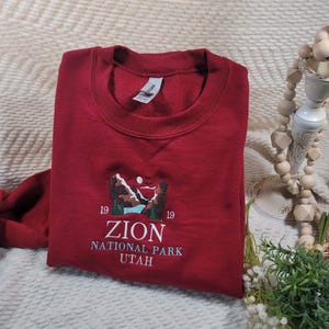 Pode incluir: Camisola vermelha com um desenho bordado de uma paisagem montanhosa e o texto "19 ZION NATIONAL PARK UTAH 19".