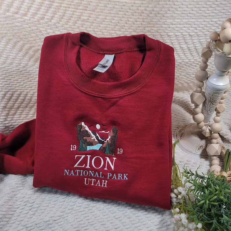 Pode incluir: Camisola vermelha com um desenho bordado de uma paisagem montanhosa e o texto "19 ZION NATIONAL PARK UTAH 19".
