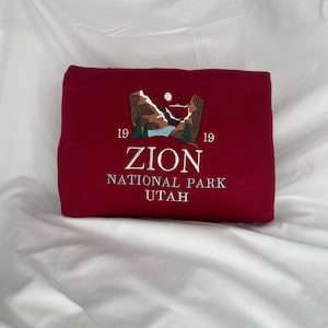 Pode incluir: Uma camisola de cor vinho com um desenho bordado de uma cordilheira com um lago e o texto "ZION NATIONAL PARK UTAH" e os n&uacute;meros "1919" de cada lado do texto.