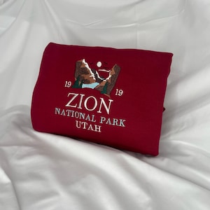 Pode incluir: Uma camisola cor de vinho com um desenho bordado de uma cordilheira com o texto "Zion National Park Utah" e os n&uacute;meros "19 19" de cada lado do texto.