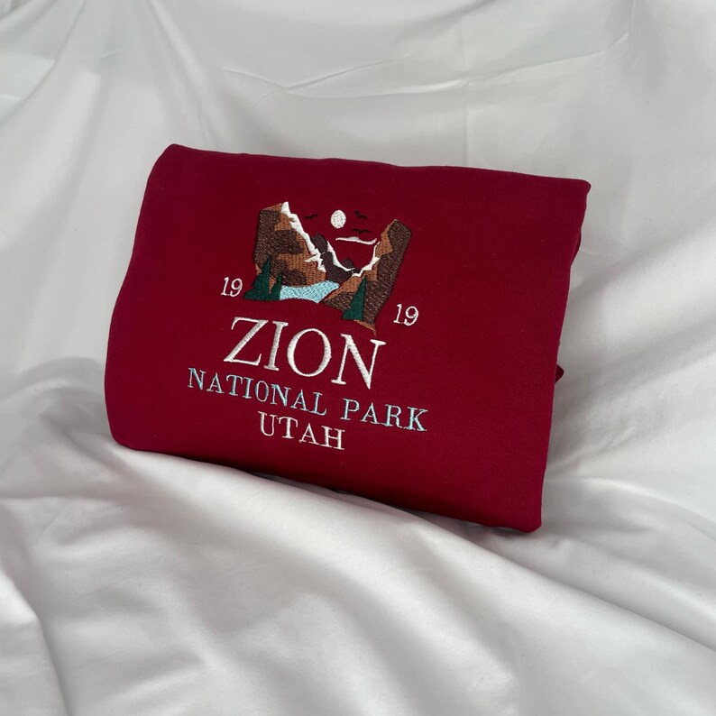 Pode incluir: Uma camisola cor de vinho com um desenho bordado de uma cordilheira com o texto "Zion National Park Utah" e os n&uacute;meros "19 19" de cada lado do texto.
