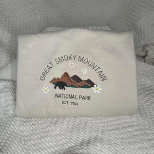 Könnte beinhalten: Ein cremefarbener Pullover mit einem gestickten Design aus einer Bergkette, einem Bären und Blumen. Der Text "Great Smoky Mountain National Park Est 1934" ist unter dem Design gestickt.