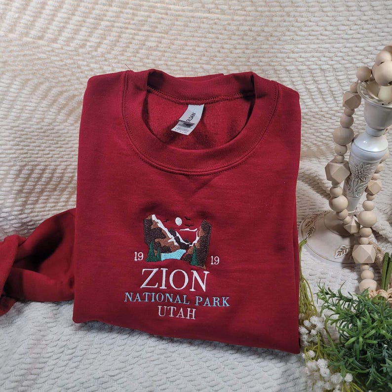 Pode incluir: Camisola vermelha com uma imagem bordada de uma paisagem montanhosa e o texto "Zion National Park Utah" com os n&uacute;meros "19" de cada lado do texto.