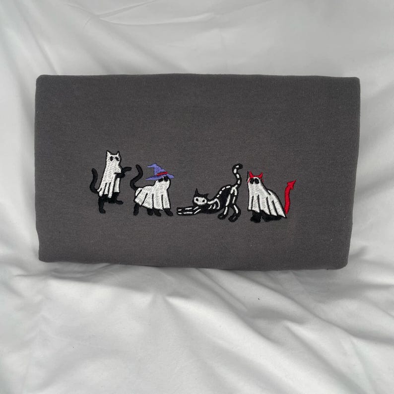May include: A gray sweatshirt with an embroidered design of five cats dressed as ghosts for Halloween. One cat is wearing a witch's hat, another has a devil's tail, and the other three are wearing simple ghost costumes.