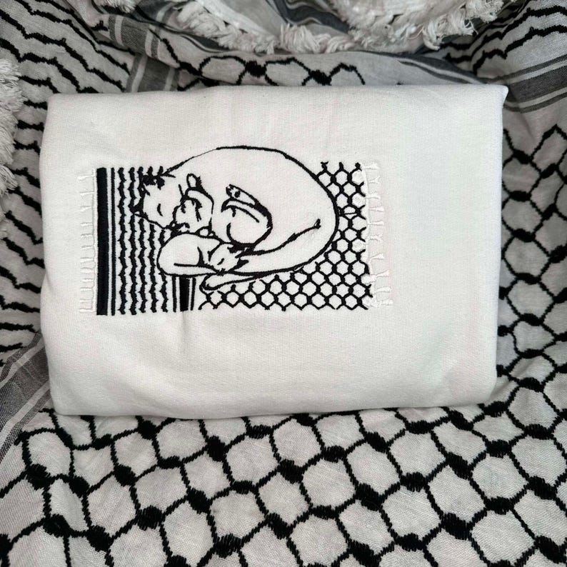 May include: White t-shirt featuring a black embroidered design of two cats cuddling. The design is framed by a black and white grid pattern.
