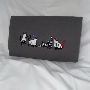 May include: A gray sweatshirt with an embroidered design of five cats dressed as ghosts for Halloween. The cats are in a line, and each one has a different costume. One cat is wearing a witch's hat, another is wearing a devil's horns, and the others are wearing simple ghost costumes.