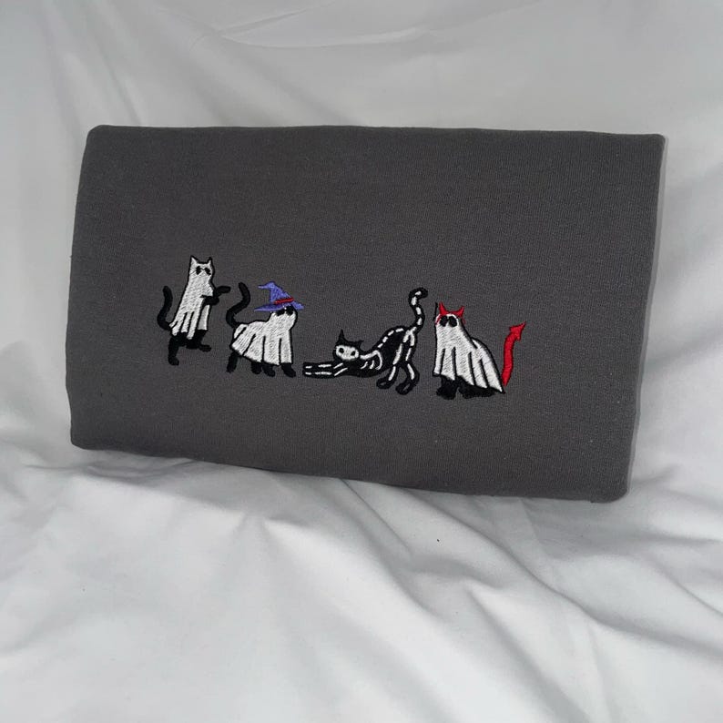 May include: A gray sweatshirt with an embroidered design of five cats dressed as ghosts for Halloween. The cats are in a line, and each one has a different costume. One cat is wearing a witch's hat, another is wearing a devil's horns, and the others are wearing simple ghost costumes.