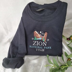 Pode incluir: Camisola de moletom preta com um desenho bordado de uma paisagem montanhosa com um rio e o texto "Zion National Park Utah" e os n&uacute;meros "1919" de cada lado.