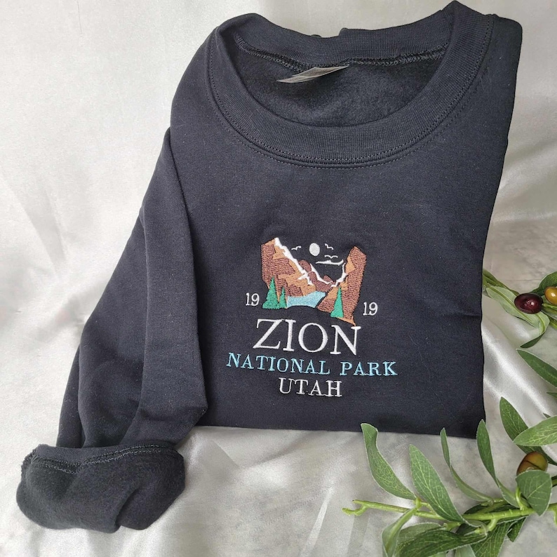 Pode incluir: Camisola de moletom preta com um desenho bordado de uma paisagem montanhosa com um rio e o texto "Zion National Park Utah" e os n&uacute;meros "1919" de cada lado.